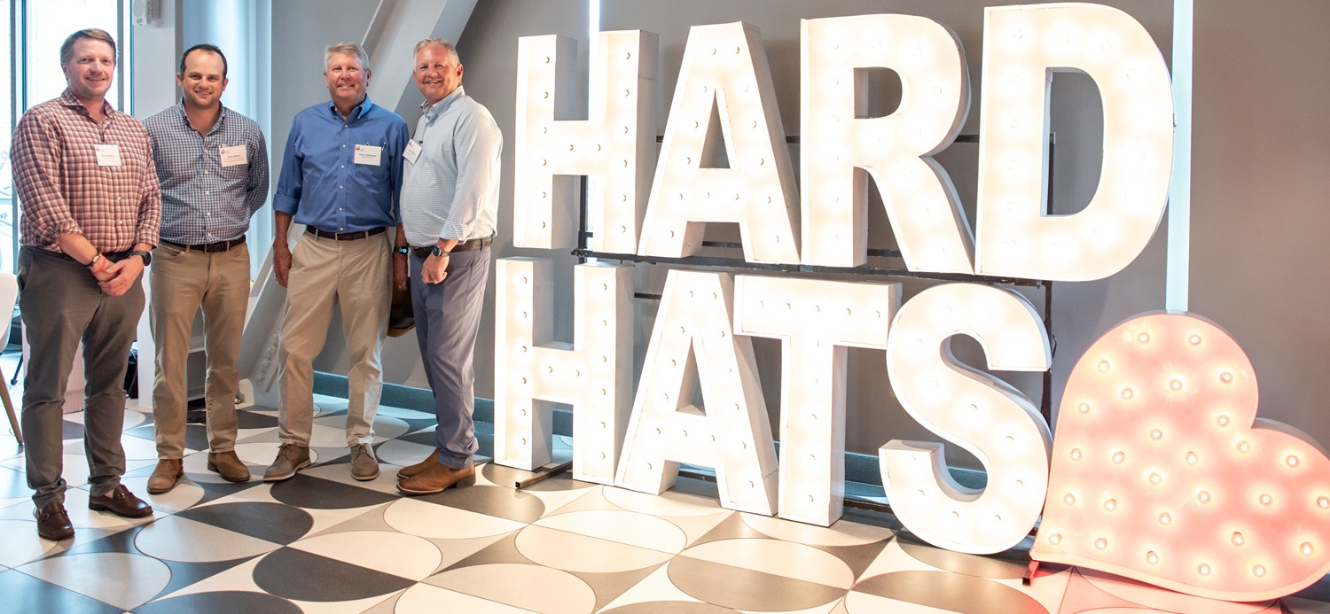 Four men in business casual attire standing next to large letters spelling out Hard Hats.